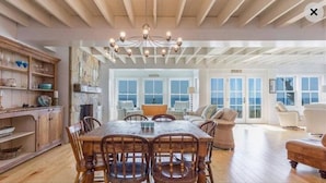 A spacious dining area with a wooden table and chairs, a chandelier, and a view of the ocean through large windows. - Beachfront Property on Private Moody Beach (Wells)