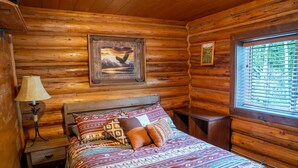 A log cabin room with a bed, a nightstand, a lamp, a window with blinds, and a framed picture on the wall.