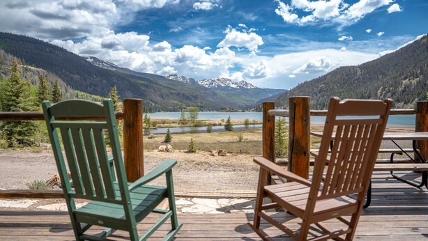 Two rocking chairs on a wooden deck overlooking a lake with mountains in the background.