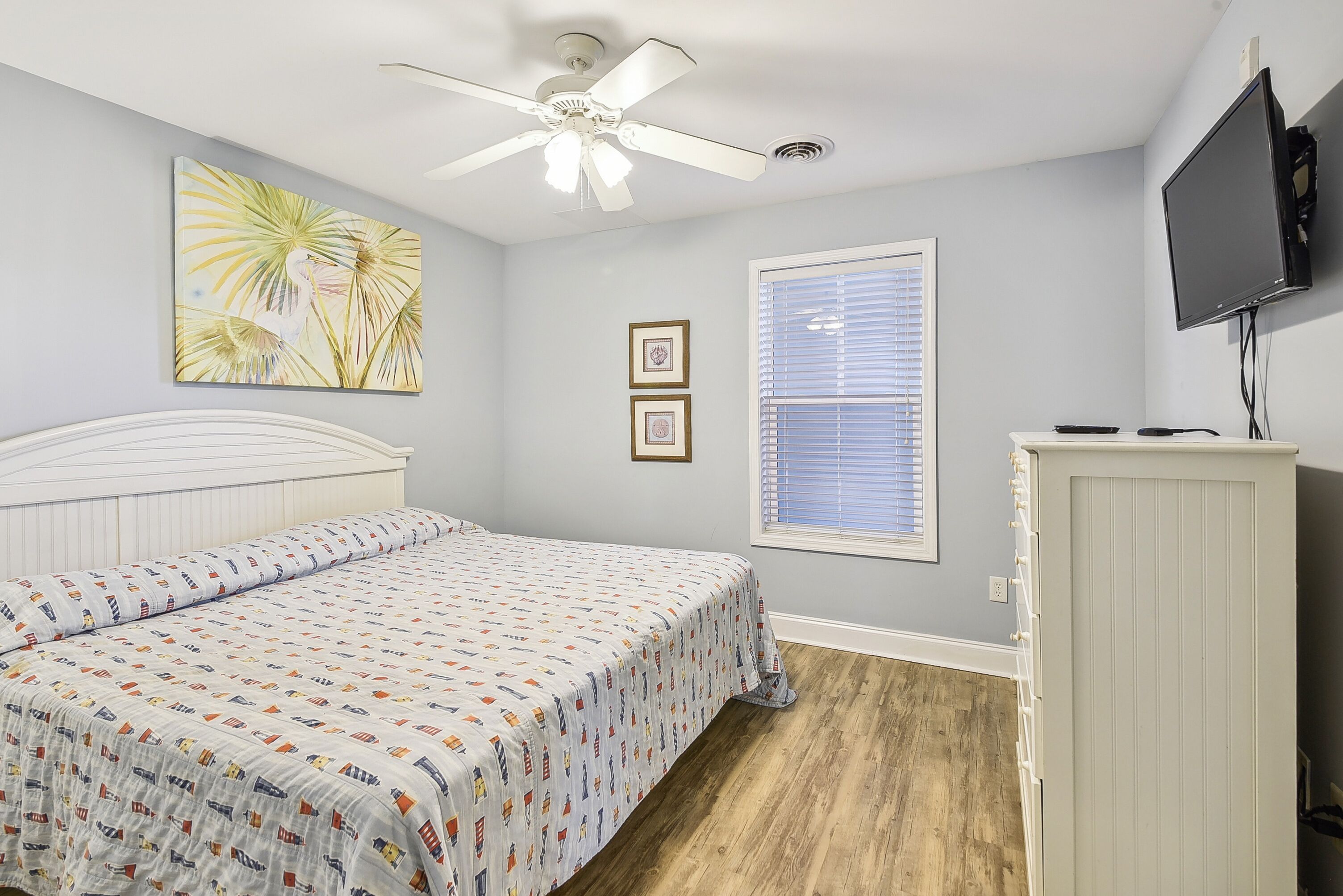 A bedroom with a bed, ceiling fan, wall-mounted TV, window, and framed pictures.