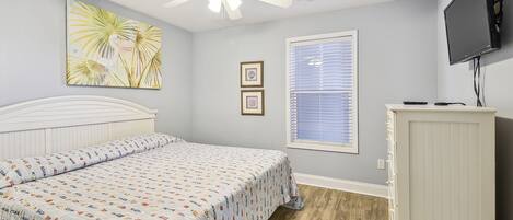A bedroom with a bed, ceiling fan, wall-mounted TV, window, and framed pictures.