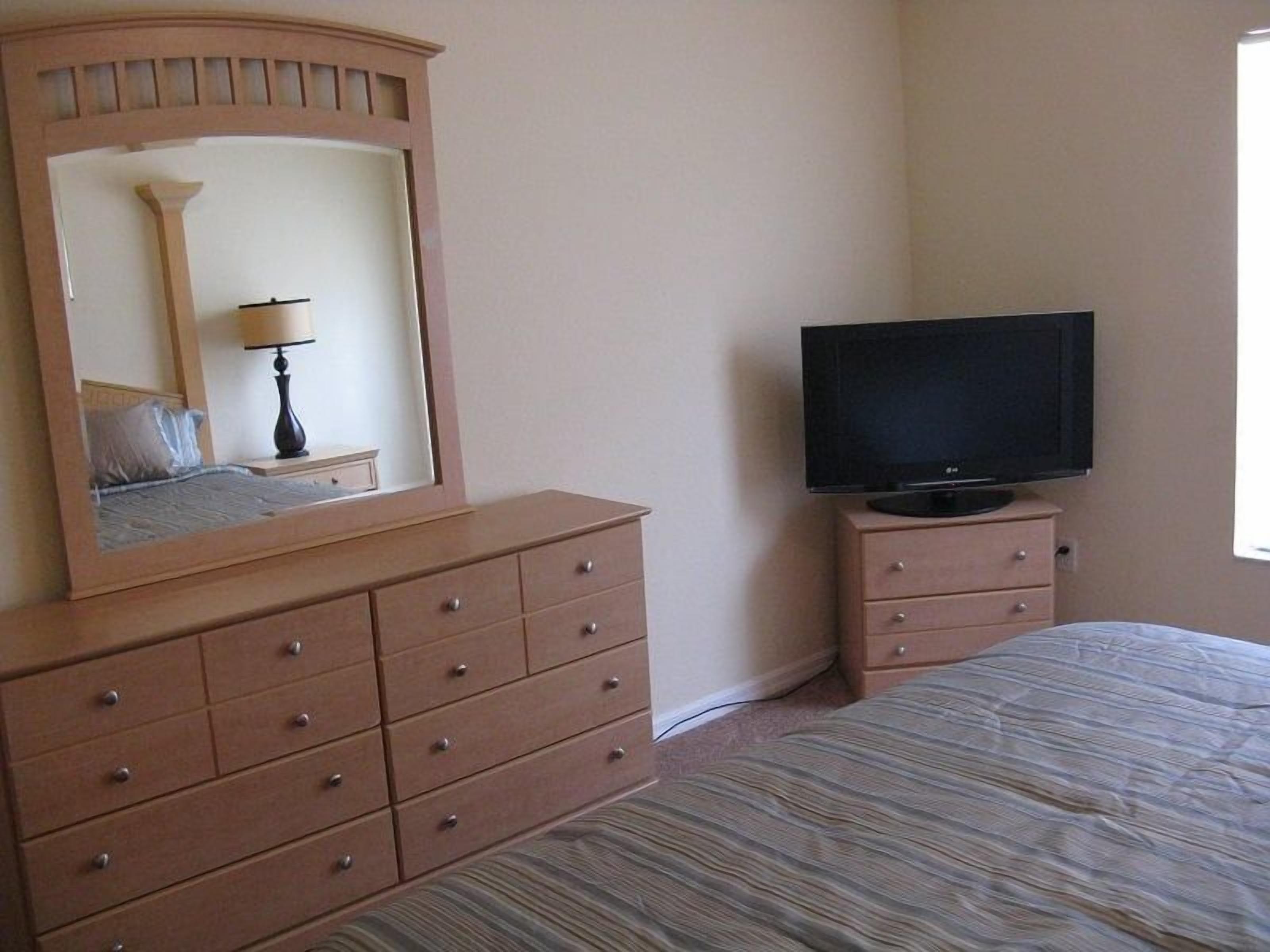 Un dormitorio con cama, cómoda con cajones, televisor sobre un mueble bajo y espejo sobre la cómoda.