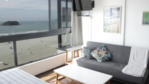 Un salón moderno con un sofá gris, una mesa blanca y vistas a la playa y al océano a través de amplias ventanas.