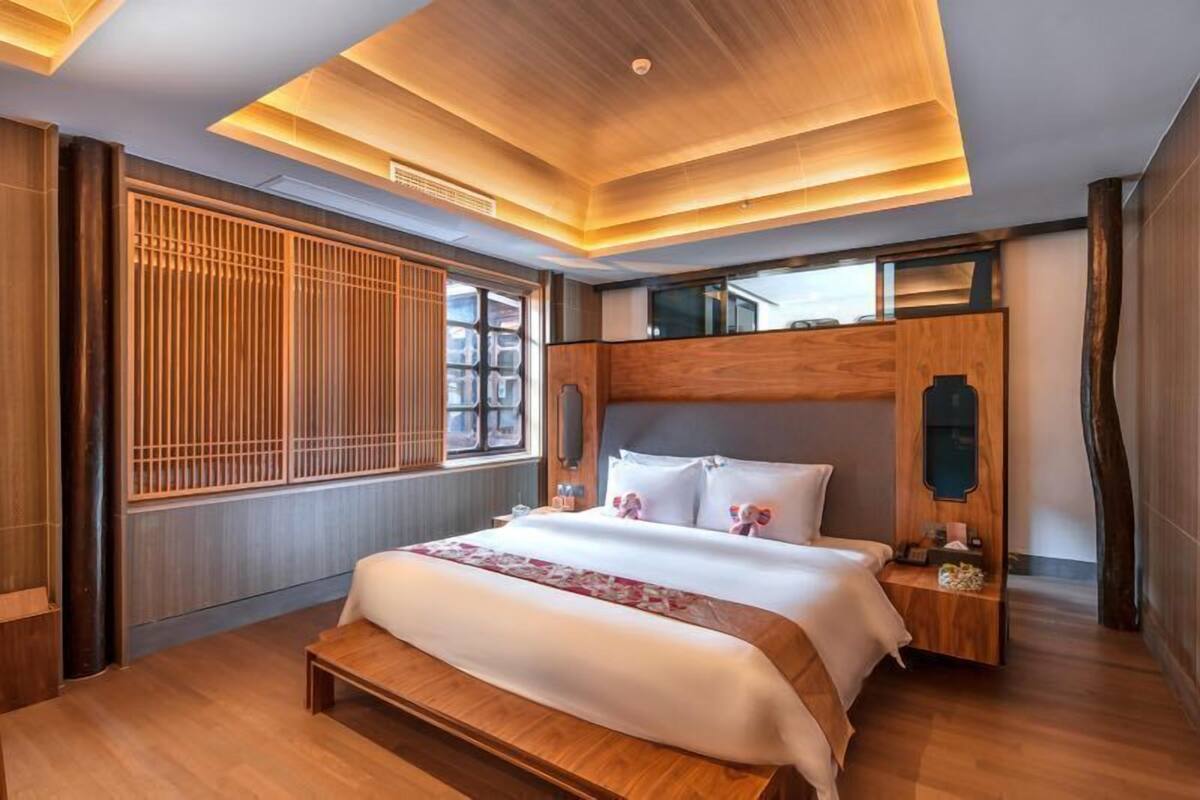 Ein modernes Hotelzimmer mit einem hölzernen Bett, einem Nachttisch, einem großen Fenster mit vertikalen Jalousien und einer Holzzellentierung an der Decke.