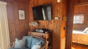 Een gezellige kamer met een aan de muur bevestigde televisie, een rieten stoel met kussens, een klein tafeltje met elektronische apparaten en een bed met een gewatteerde spreidek.
