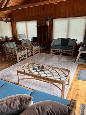 Interior acogedor de una cabaña de madera con mobiliario de mimbre, una mesa de centro de vidrio y un sofá azul.