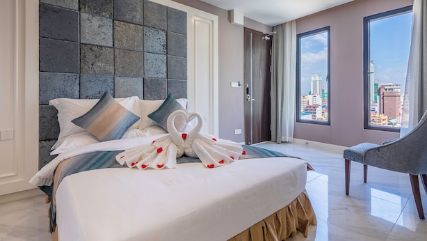 Quarto de hotel com uma cama grande, cabeceira moderna, vista da cidade através de amplas janelas e um espaço de estar com uma poltrona.