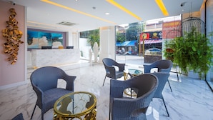 Um saguão de hotel com balcão de recepção, área de estar com cadeiras de vime e uma mesa de vidro, e vista para a rua.