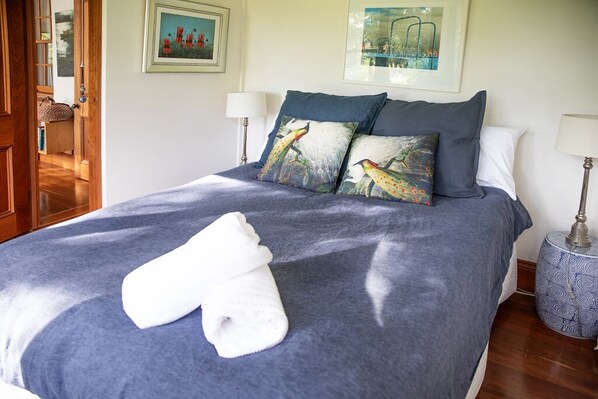 Cama bem arrumada com manta azul e duas almofadas, toalha branca sobre a cama, quadro na parede e luminária na mesa de cabeceira.