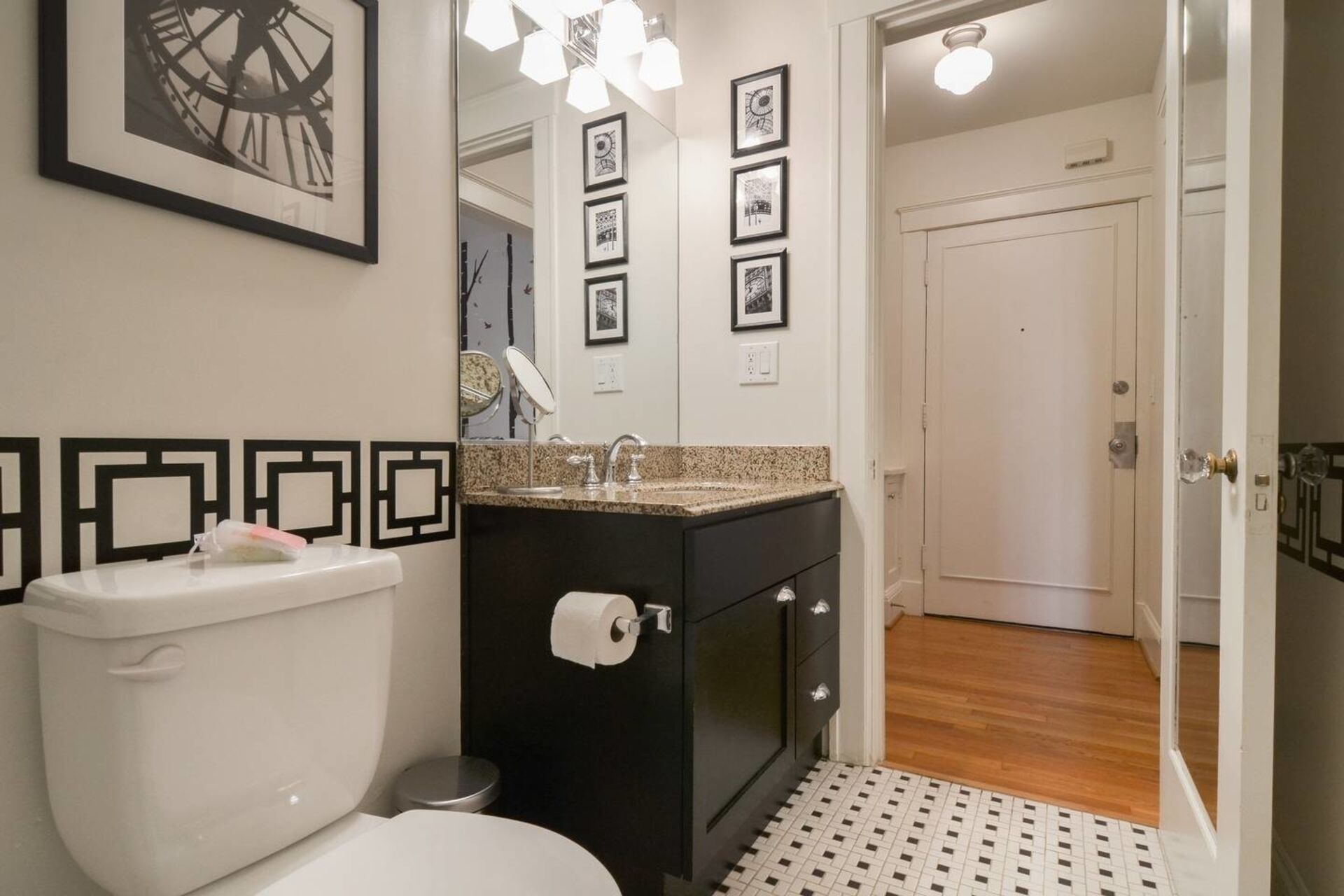 A bathroom with a toilet, sink, and mirror. There are framed pictures on the wall and a door leading to another room.