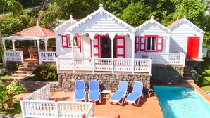 En vit byggnad med röda detaljer, en swimmingpool och blå solstolar.