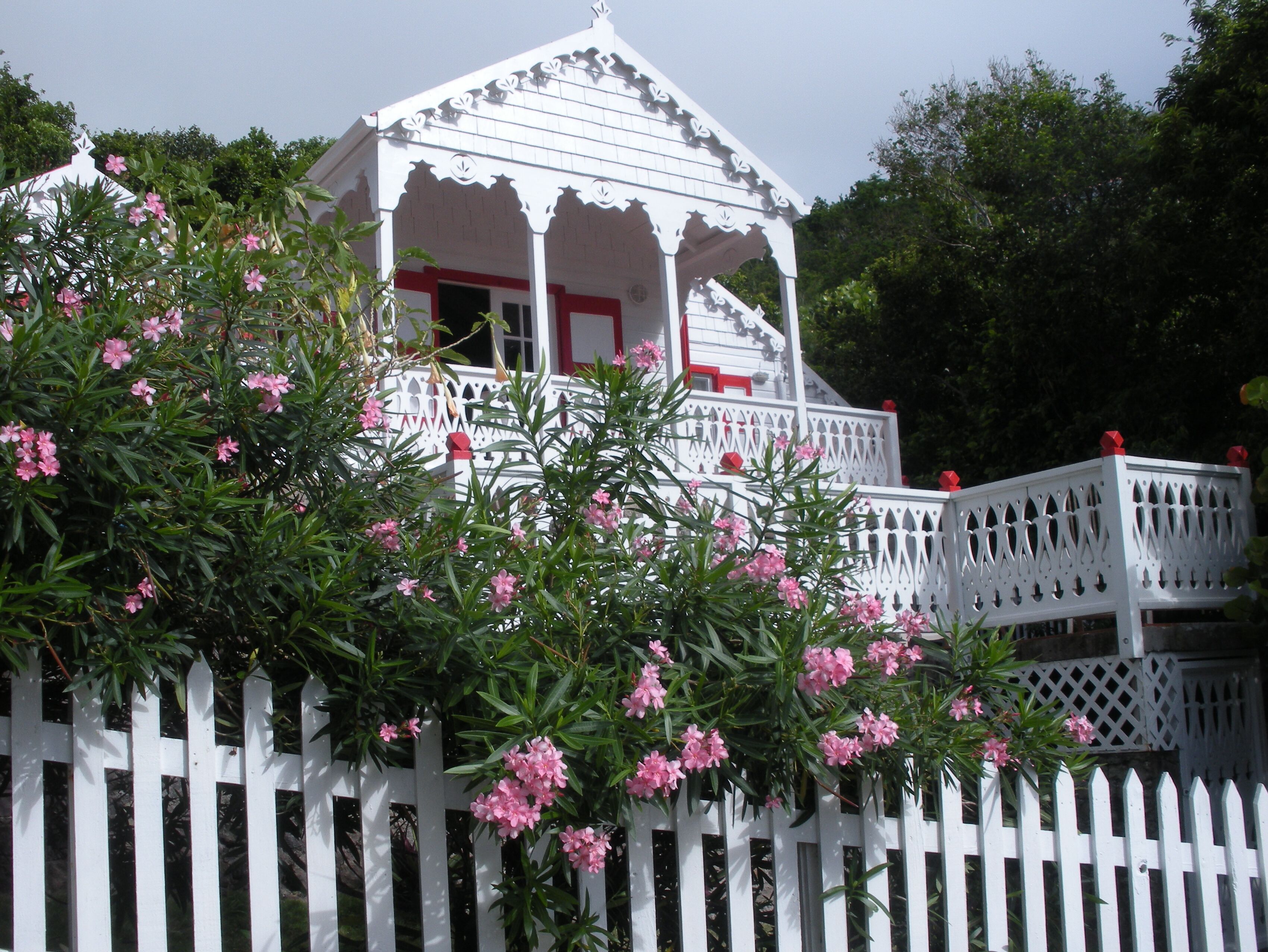 Een witte gazebo met een rode deur, omringd door een witte picket hek en roze bloeiende planten.