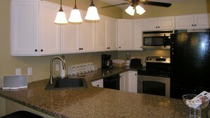 Cozinha com armários brancos, bancada de granito, geladeira preta, micro-ondas, fogão, ventilador de teto e luminárias pendentes.