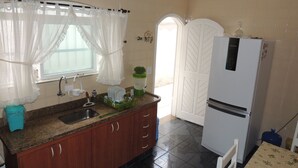 A kitchen with a sink, cabinets, a refrigerator, and a window with curtains. - House 1 Suite and 2 Bedrooms - Jd. Suarão - Itanhaém (Itanhaém)