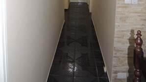 A narrow hallway with a tiled floor, a wooden door at the end, and a light fixture overhead. - House 1 Suite and 2 Bedrooms - Jd. Suarão - Itanhaém (Itanhaém)