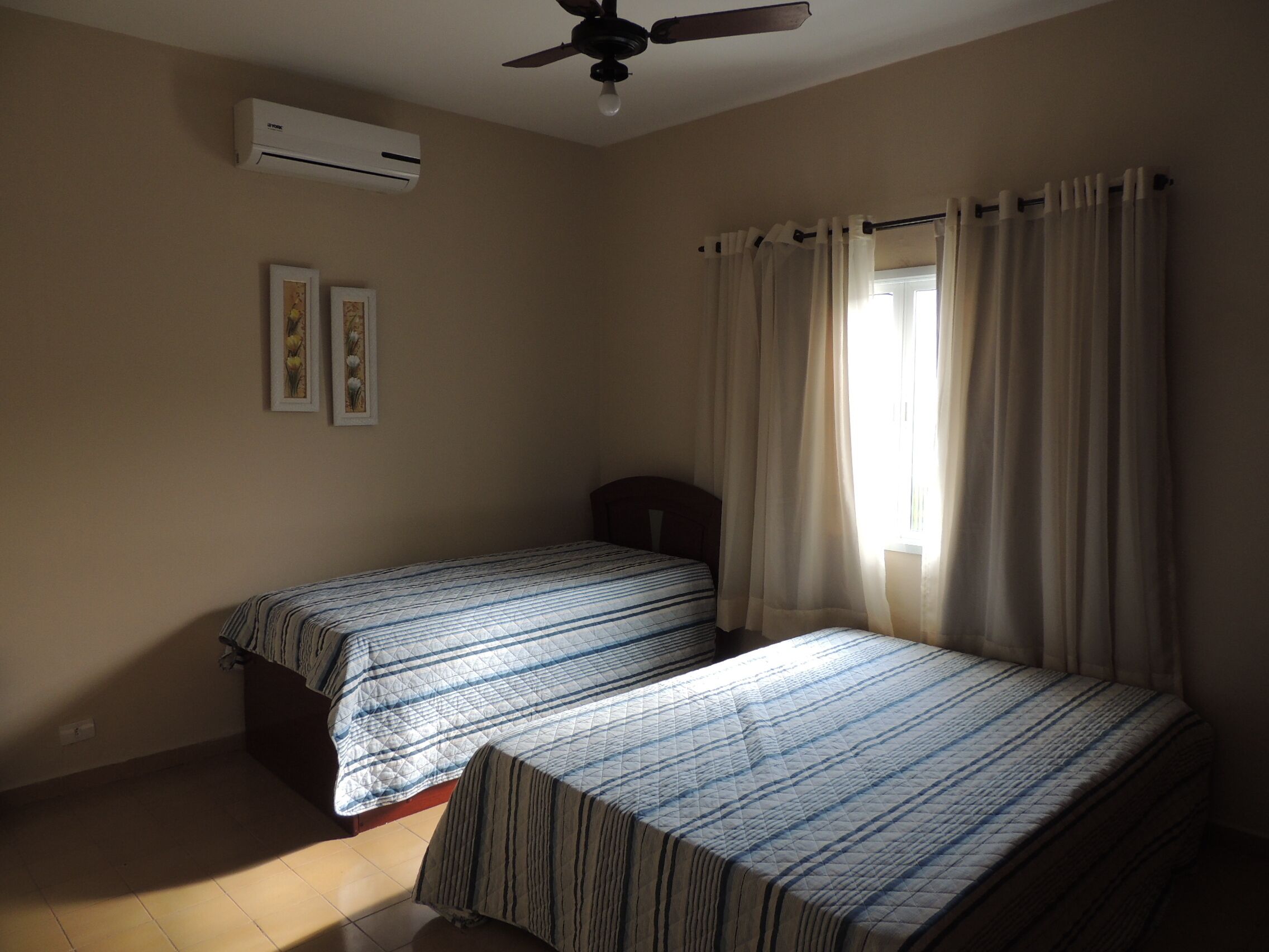 Un dormitorio con dos camas, ventilador de techo, aire acondicionado y ventana con cortinas.