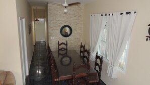 A dining area with a long table, chairs, a ceiling fan, a mirror, and a window with curtains. - House 1 Suite and 2 Bedrooms - Jd. Suarão - Itanhaém (Itanhaém)