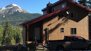 Une maison en bois à deux étages, avec un toit rouge, un porche à l’avant et un camion garé devant. On aperçoit, au loin, une montagne dont le sommet est enneigé.