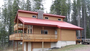 Une cabane à deux étages avec un toit rouge, un balcon à l’étage supérieur et un garage attenant au bâtiment principal.