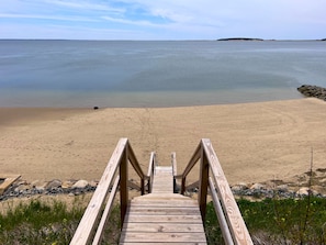 A wooden boardwalk leading to a sandy beach with calm water and a distant landmass.