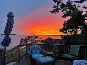 A deck with a wicker chair and a small table, overlooking a body of water with a sunset.
