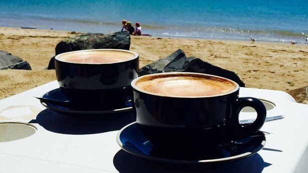 Deux tasses de café noir sur une table blanche, avec une vue sur la plage en arrière-plan.