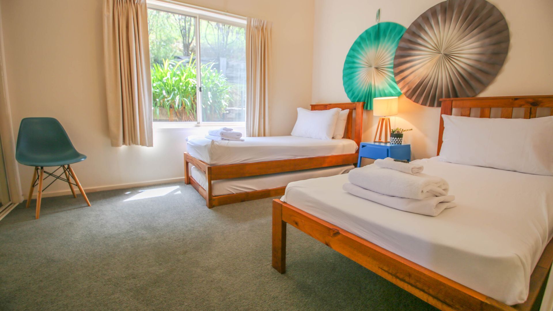 A hotel room with two single beds, a blue chair, a bedside table with a lamp, and a view of greenery through the window.