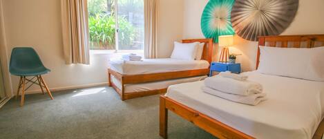 A hotel room with two single beds, a blue chair, a bedside table with a lamp, and a view of greenery through the window.