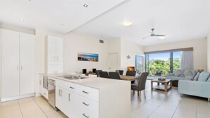 A modern kitchen with white cabinetry, a dining area with a table and chairs, and a living area with a sofa and a view of greenery.