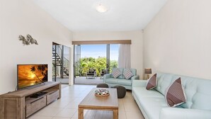 A living room with a TV, a wooden coffee table, a light blue sofa, and a balcony with a view of greenery.