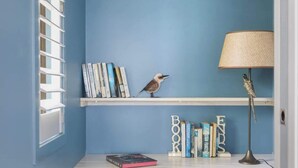 Ein Zimmer mit blauer Wand, einem weißen Schreibtisch mit Büchern und einer Vogelfigur, einer Lampe und einem Stuhl.