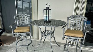 A round table with two wicker chairs, each with a cushioned seat, and a black lantern on the table. - Waterfront home with south facing pool & views over the bay (Marco Island)