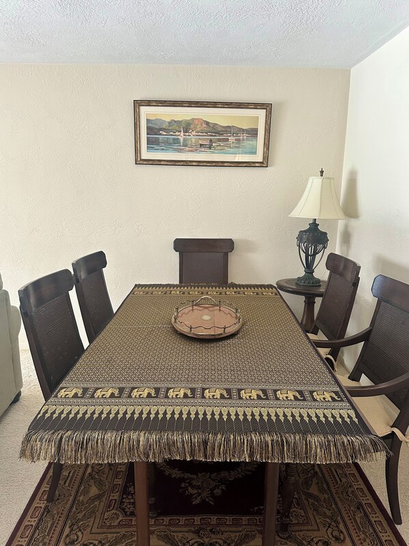 Un comedor con una mesa preparada para cuatro personas, rodeada de sillas, un cuadro en la pared y una lámpara sobre una mesita auxiliar.