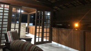 A room with a wooden ceiling, large sliding glass doors, a cushioned chair, and a small table. - Itaipu Beach House - Ocean View (Itaipu)