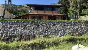 A two-story house with a stone wall in front, a balcony, and a view of a hill. - Itaipu Beach House - Ocean View (Itaipu)