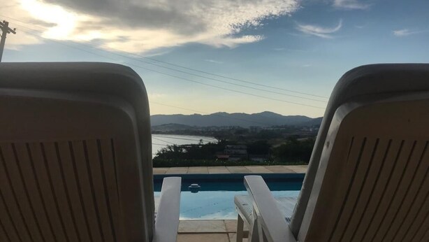 Two lounge chairs facing a swimming pool with a view of a lake and mountains in the distance. - Itaipu Beach House - Ocean View (Itaipu)