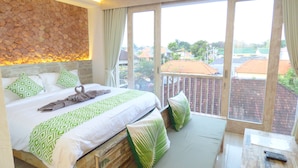 Un dormitorio con una cama grande, una pared de piedra, un balcón con vistas a edificios y una mesa de madera con un control remoto.