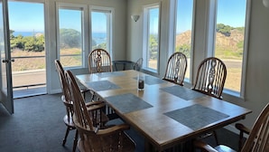 A dining area with a large table and chairs, large windows with a view of the outdoors, and a door leading outside. - All About the View North 3 bedroom oceanfront (Bandon)