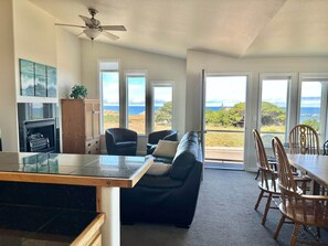 A spacious living area with a fireplace, a sofa, a dining table with chairs, and large windows offering a view of the ocean.