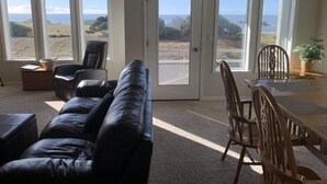 A living room with a black leather couch, a wooden dining table with chairs, and large windows offering a view of the landscape. - All About the View North 3 bedroom oceanfront (Bandon)