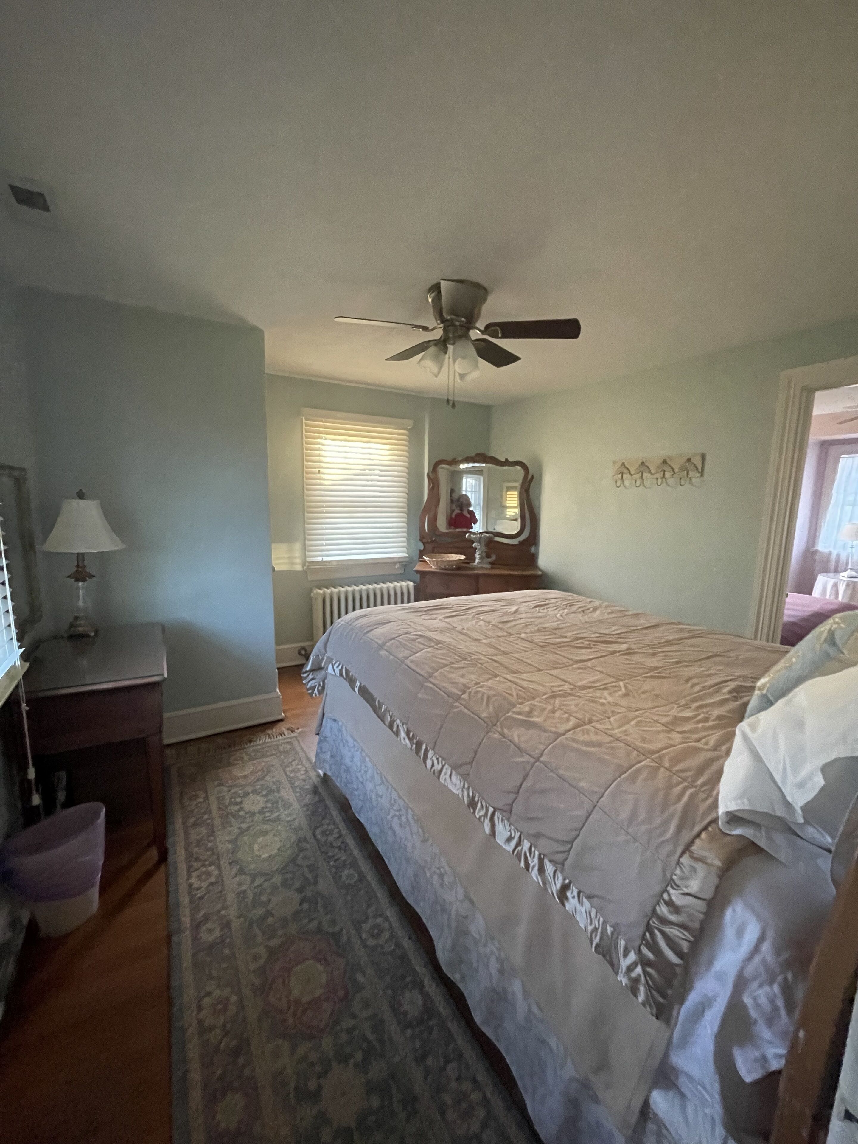 A bedroom with a bed, a ceiling fan, a dresser, a lamp, a mirror, and a window with blinds.