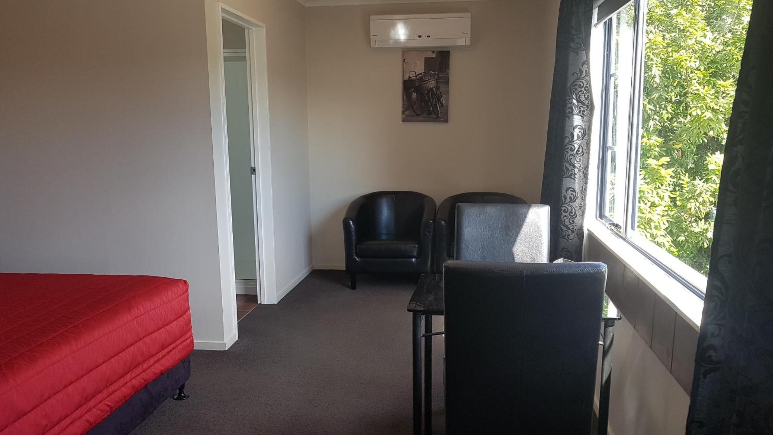 A small room with a red bed, two black chairs, a small table, and a window with a view of trees.