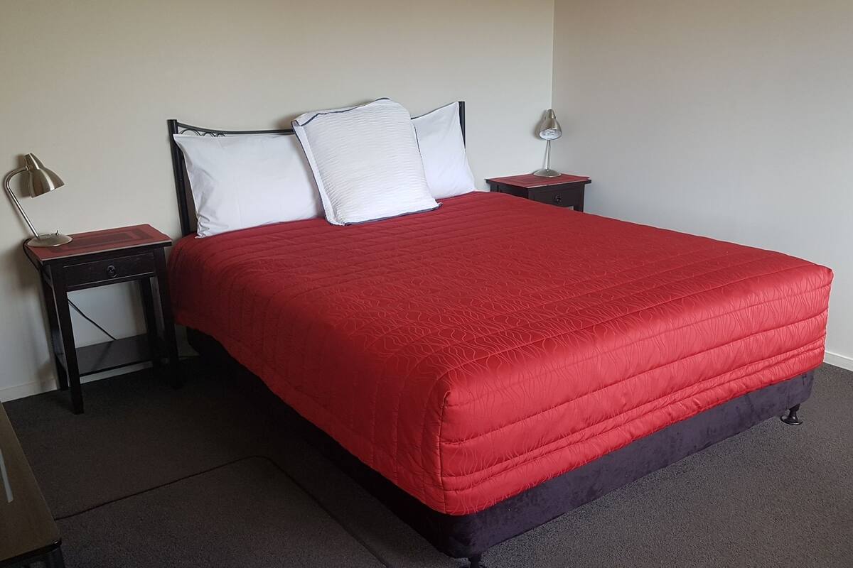 A neatly made bed with a red bedspread and white pillows, two bedside tables with lamps, and a dark floor.