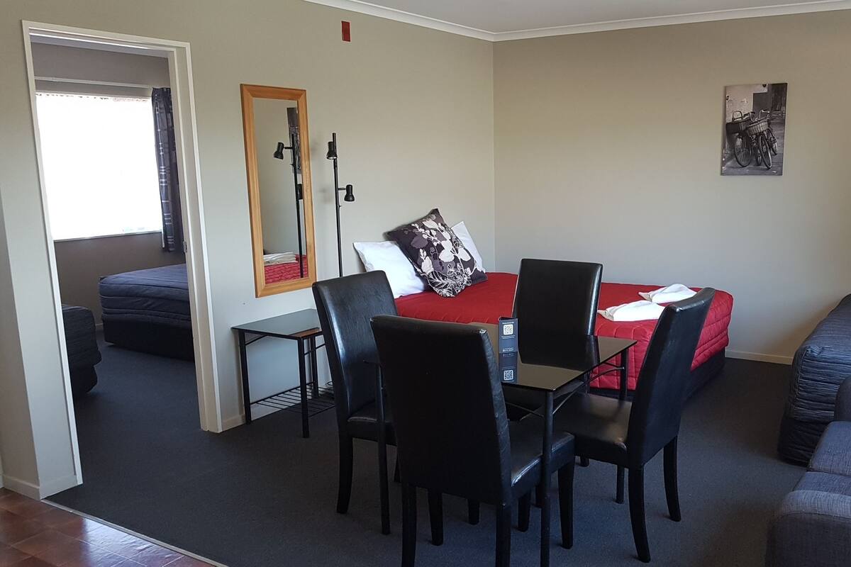 A compact hotel room with a bed, a desk with chairs, a small table, and a mirror.