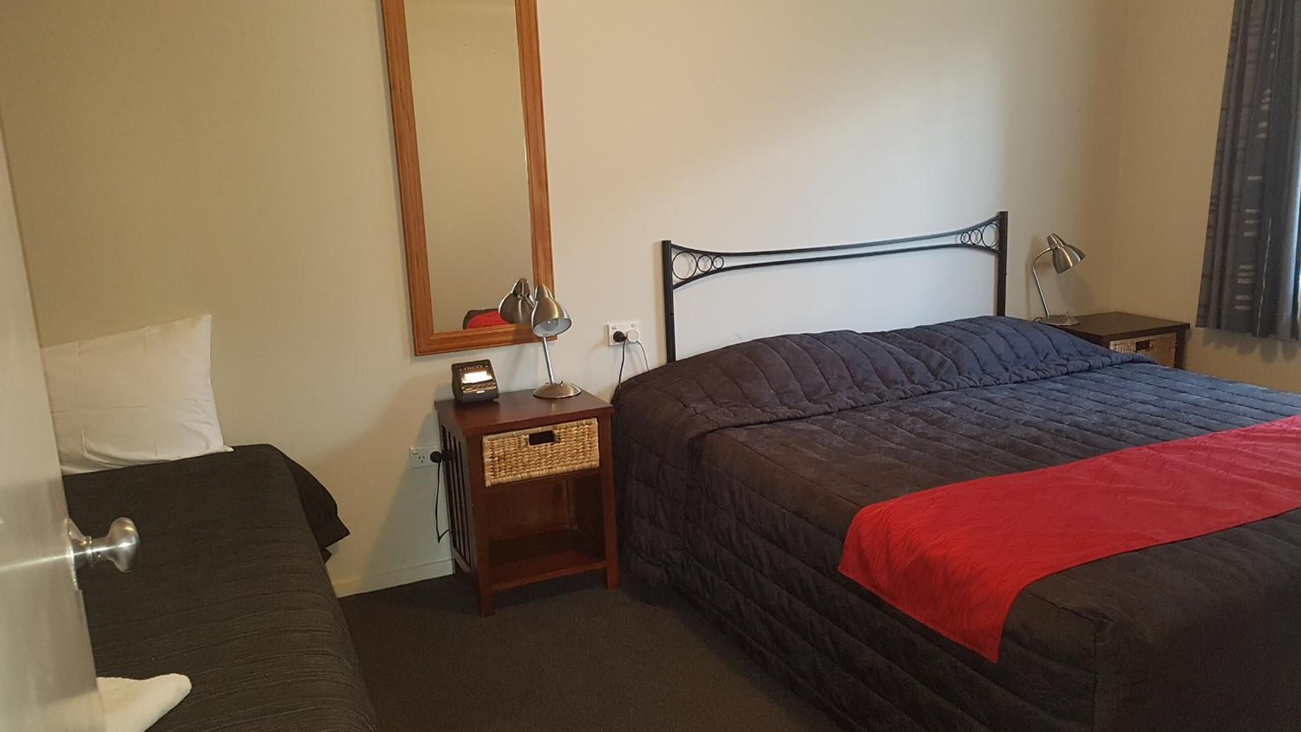 A hotel room with a bed, a bedside table with a lamp, a nightstand, a mirror, and a wall socket.
