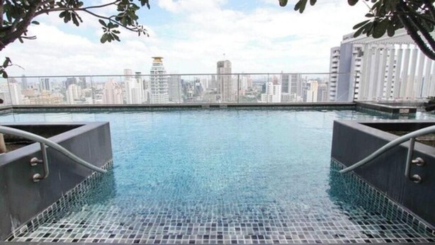 A rooftop infinity pool with a city skyline view, surrounded by a metal railing and a tiled floor.