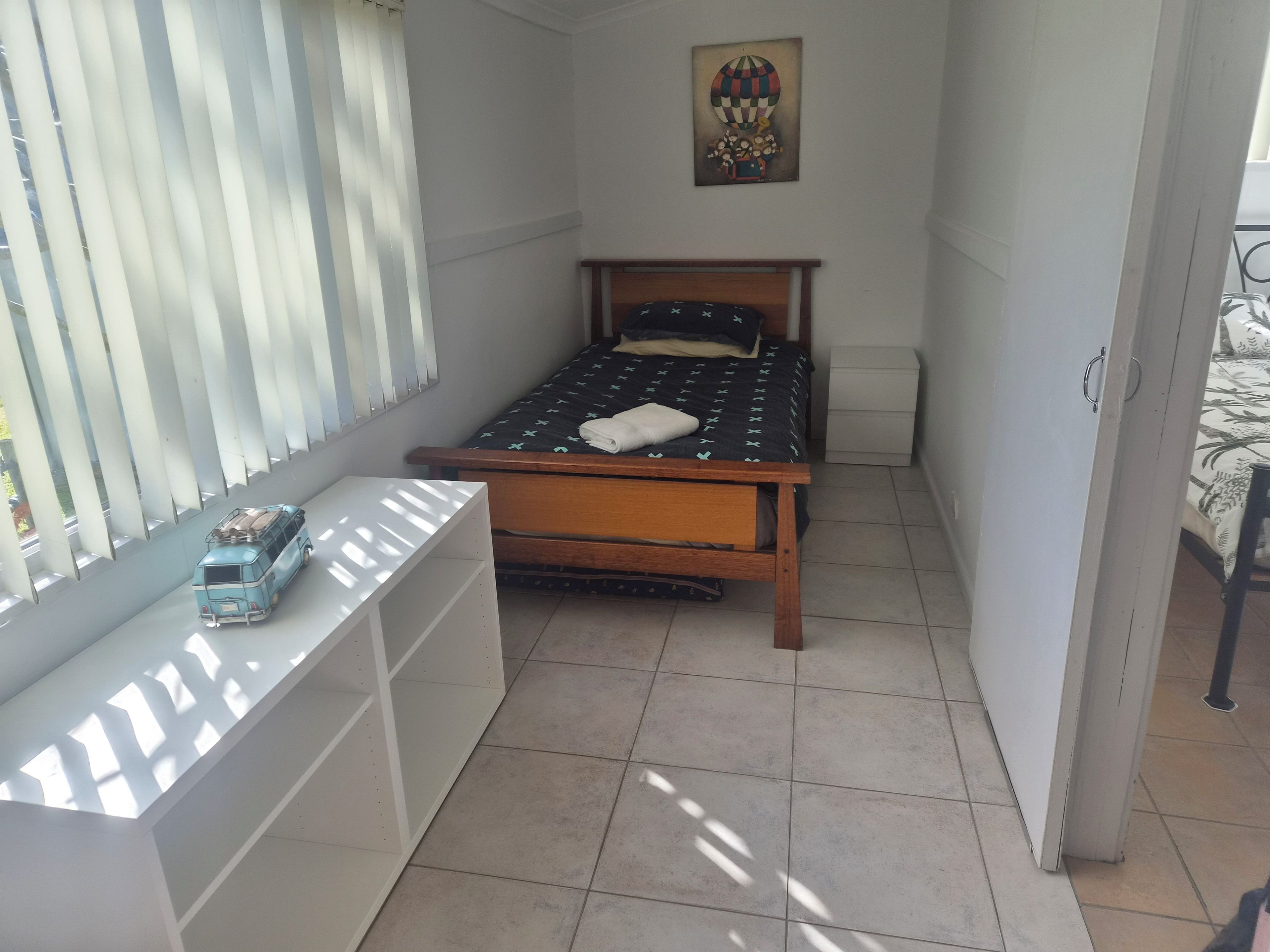 Une petite pièce étroite, avec un lit simple, une fenêtre avec des stores, une petite table et une œuvre d’art accrochée au mur.