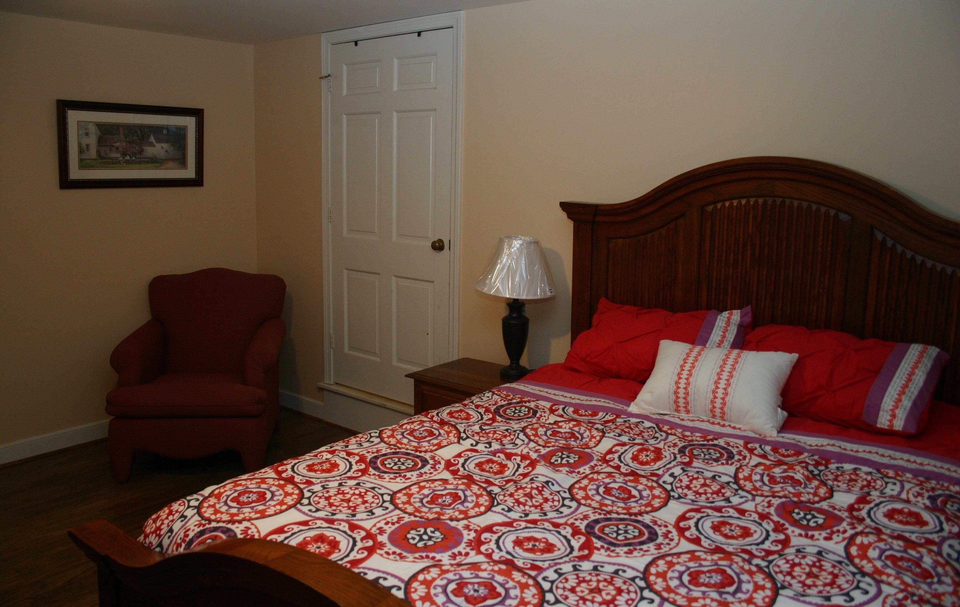 Une chambre à coucher avec une tête de lit en bois, un fauteuil rouge, une couvre-lit à motifs et une porte surmontée d’un tableau encadré.