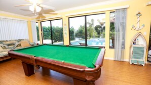 Ein Billardtisch in einem Raum mit großen Fenstern, einem Deckenventilator und Blick auf einen Swimmingpool.