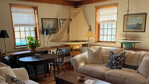 A cozy living room with a wooden ceiling, a large window with a sailboat model, a dining area with a table and chairs, and a sofa with patterned cushions. - Charming Nantucket home. (Nantucket)
